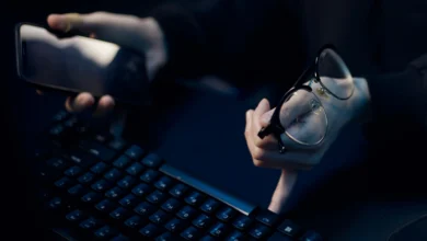 Karanlık ortamda bir elinde telefon, diğer elinde gözlük tutarak klavyeye basan kişi.