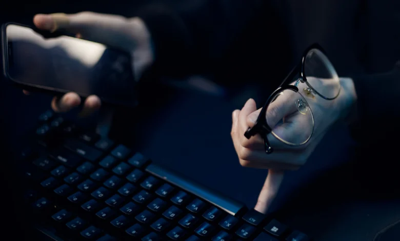 Karanlık ortamda bir elinde telefon, diğer elinde gözlük tutarak klavyeye basan kişi.